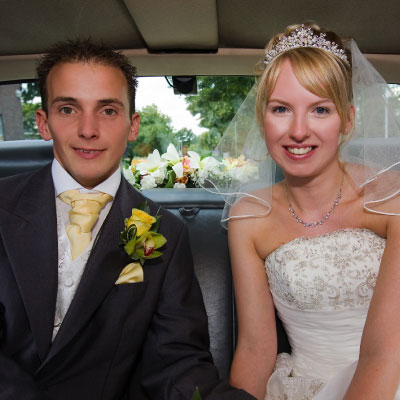Two individuals in wedding attire seated in the back of a luxury vehicle. The person on the left wears a suit with a yellow boutonniere, while the person on the right wears a white wedding dress with intricate lace details and a tiara. The backseat is adorned with flowers, suggesting this is part of a chauffeured business offering wedding car services
